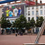 Vue lointaine de jour de face illustrant l'intégration urbaine parfaite de cette campagne grand format au cœur de Lyon, sur la place Bellecour.